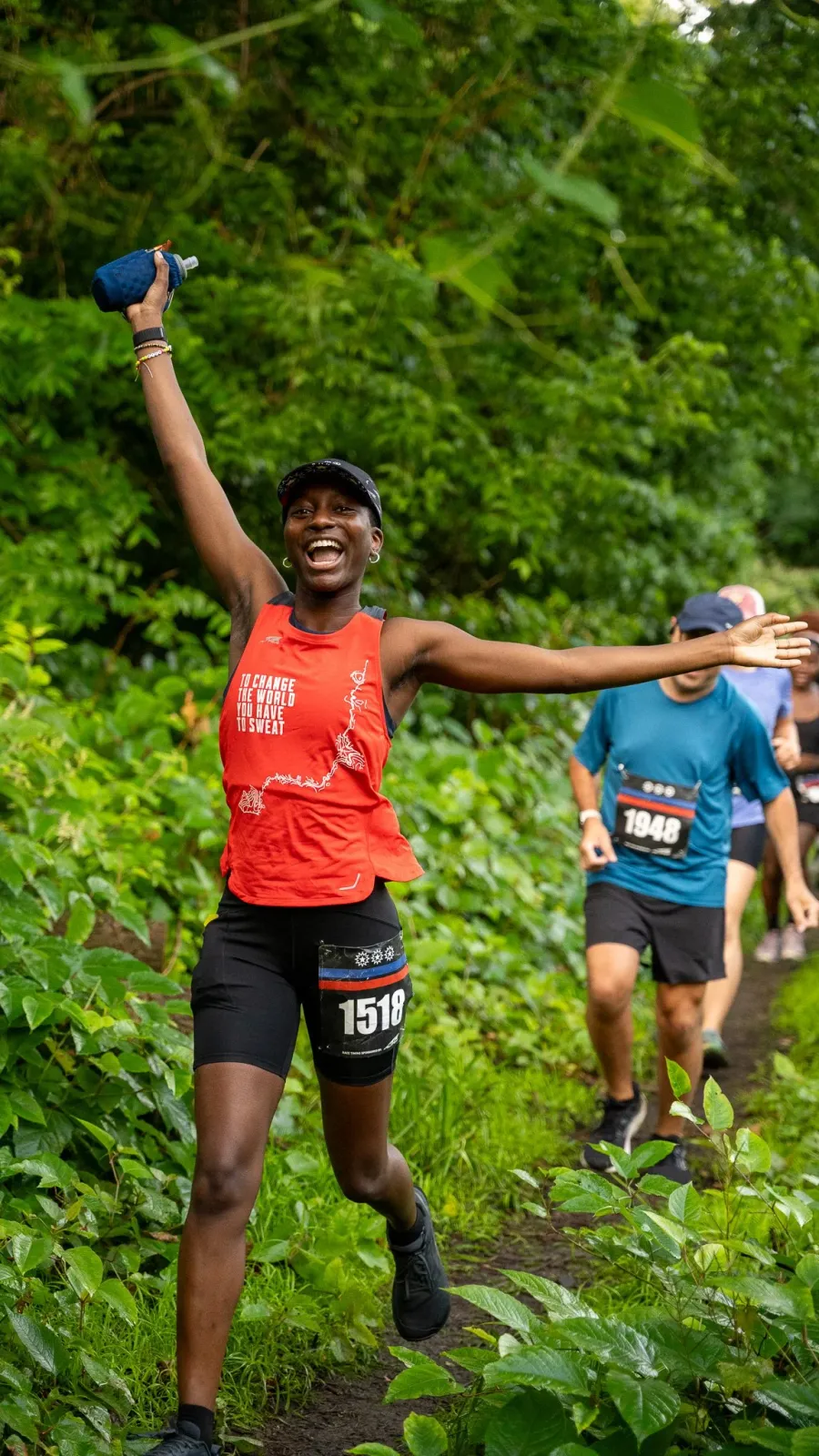 A female runner with arms outstretched and a wide smile, holding a water bottle, running on a path surrounded by greenery.