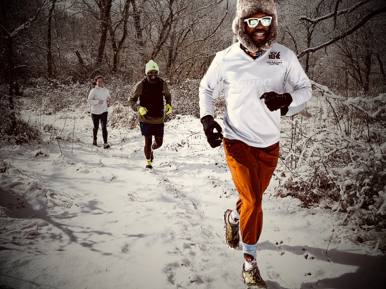 Three runners on a snowy trail through a forest, with the lead runner in the foreground wearing a white long-sleeved shirt and orange pants.