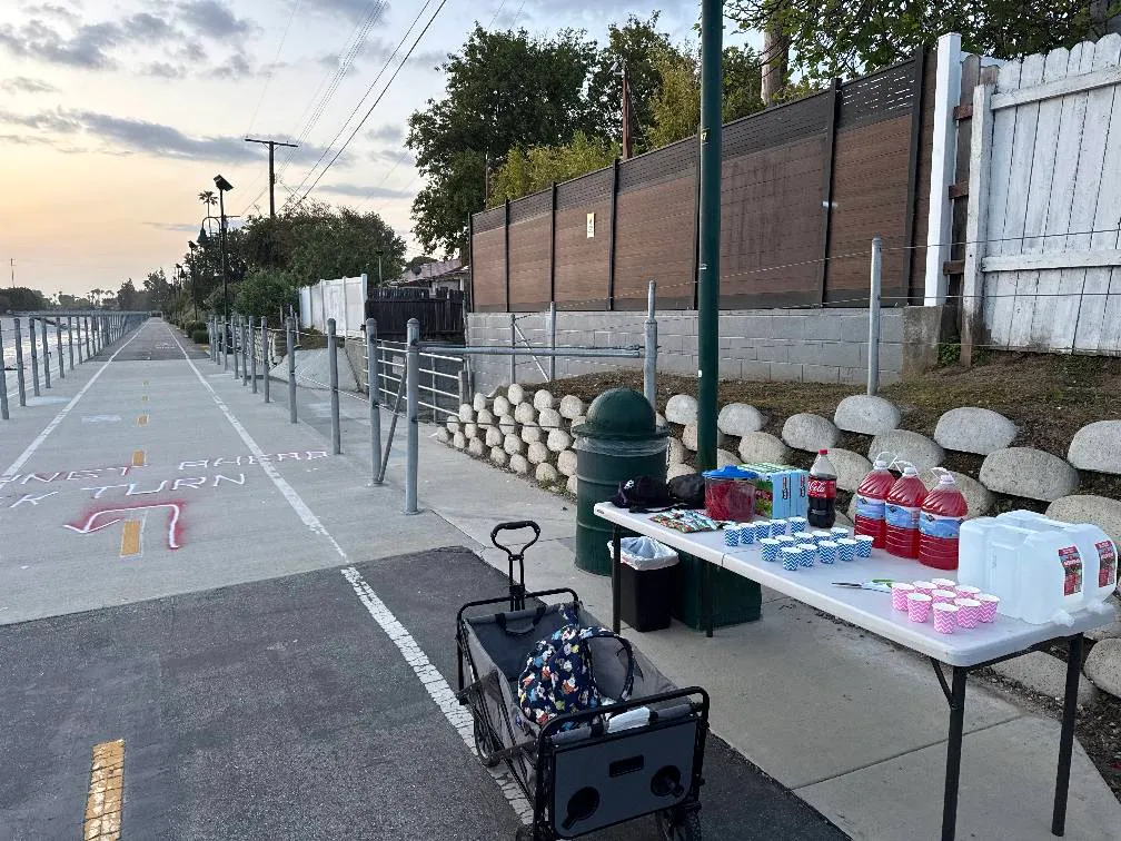 A scenic waterfront path with yellow and white lane markings, a table with drinks and cups set up for a race, and a clear sky at dusk.