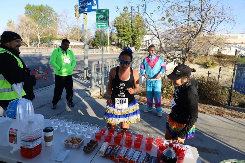 Runners and volunteers at a race aid station with drinks and snacks, with a sign for the Los Angeles River in the background.