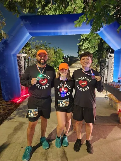 Three smiling runners, two men and one woman, stand under a blue inflatable archway, holding up their race medals. They are wearing matching black long-sleeved shirts with a red heart logo and the text "SUNNYDALE SLAYERS" and orange baseball caps.