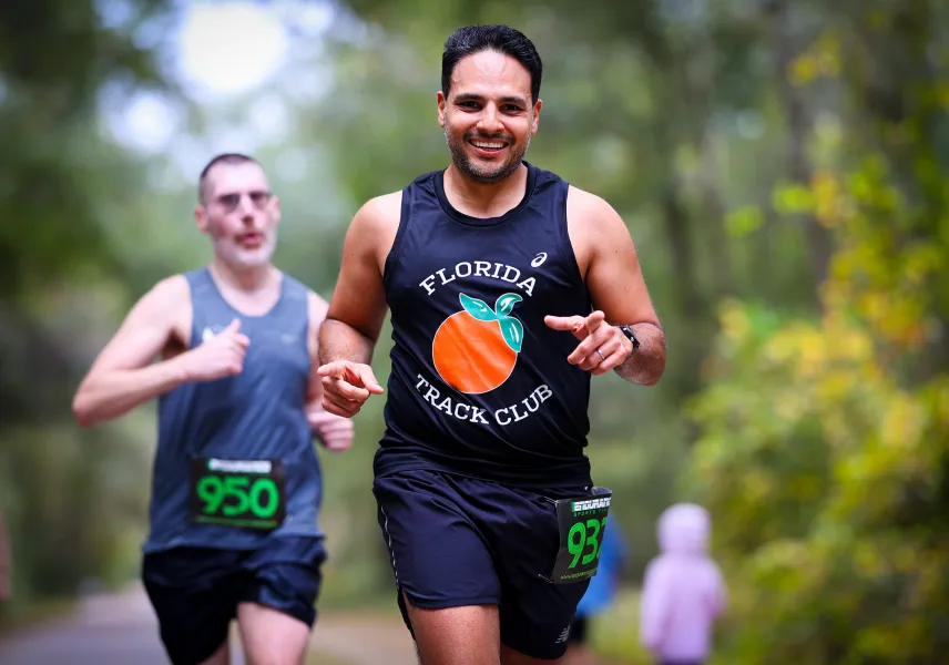 A smiling male runner in a black Florida Track Club tank top points towards the camera while running on a tree-lined path.
