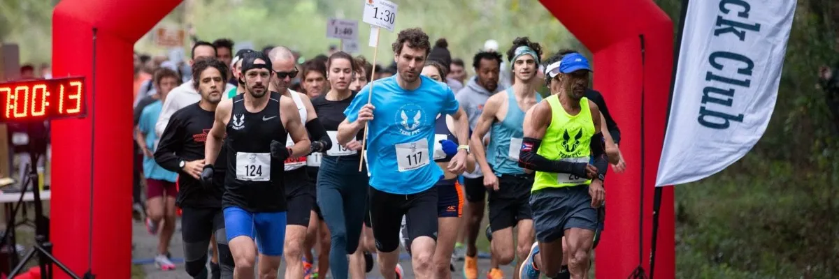 Runners starting a race, passing under a red archway with a digital clock showing 7:00:13.