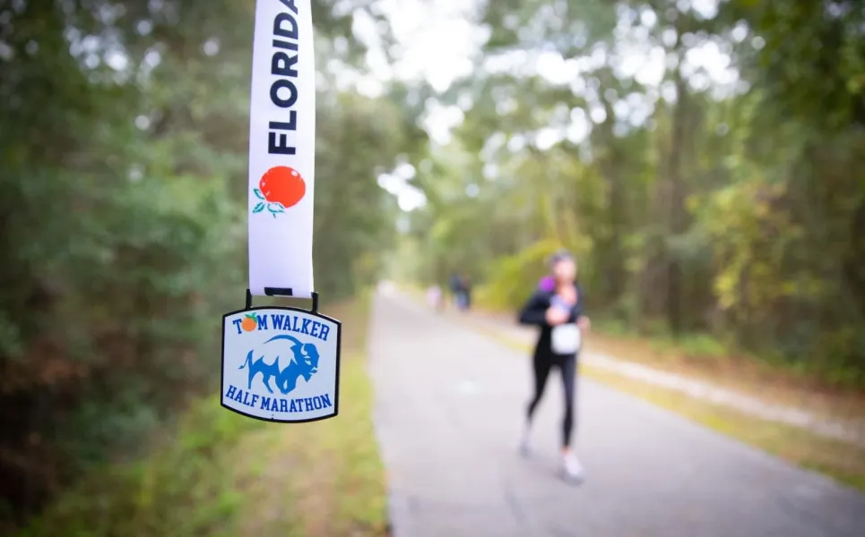 A race medal hangs in the foreground with 'Tom Walker Half Marathon' visible, while a runner is blurred in the background on a paved path surrounded by trees.