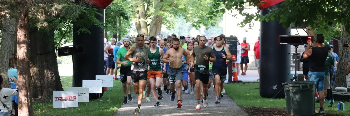 A large group of runners are lined up at the start of a race, with some in the foreground actively running forward.