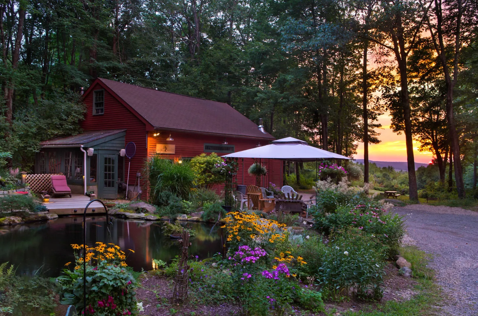 Scenic view of a red building with a pond and gardens, set against a forest backdrop at sunset.