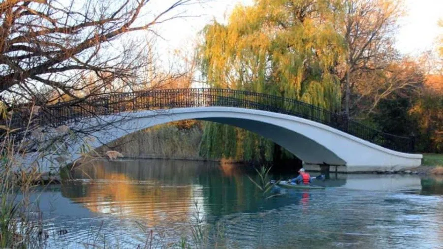 A white arched bridge over a calm body of water, with trees and foliage on the banks. A person in a kayak is visible on the water.
