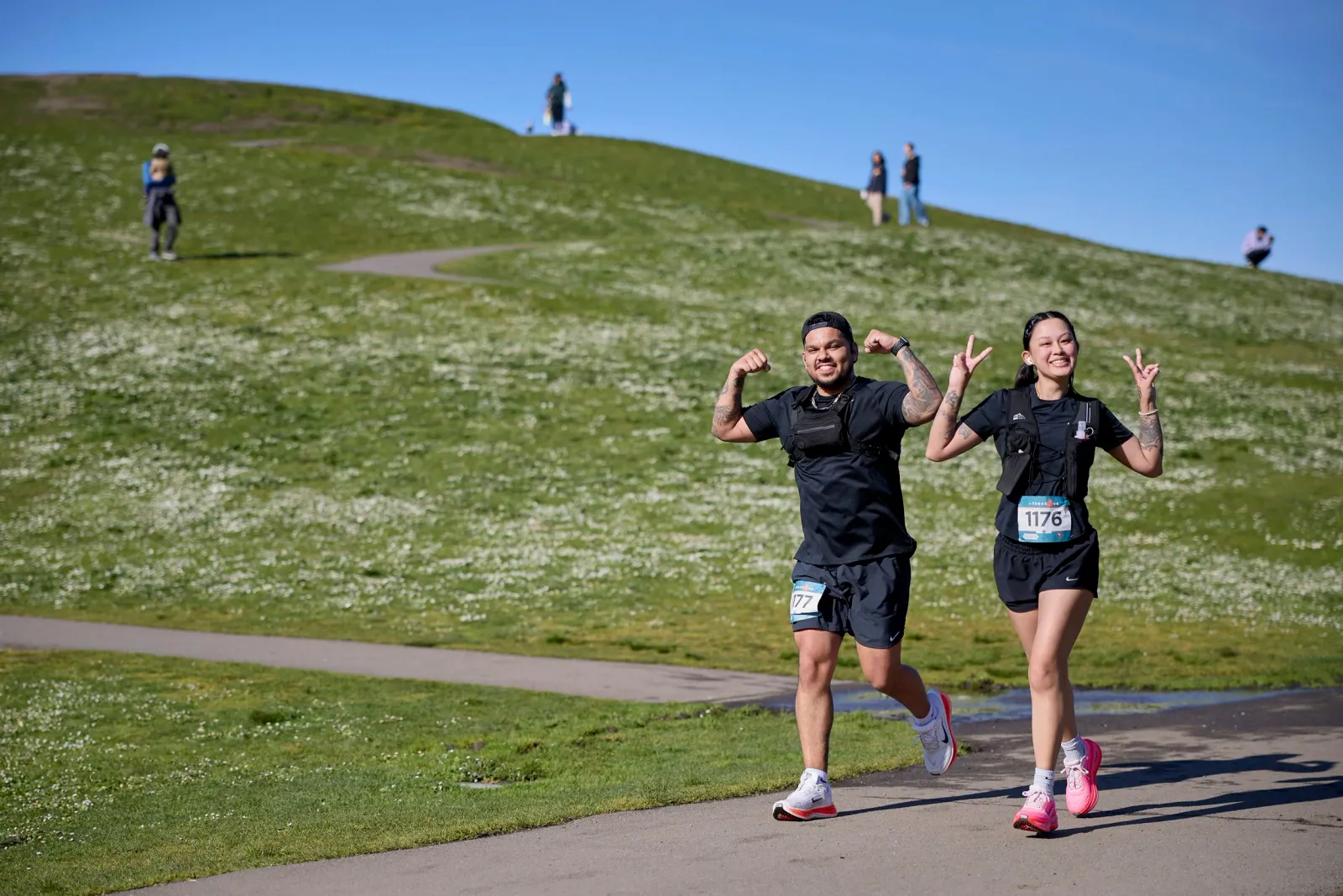 A male and female runner smile and flex their muscles while running on a paved path through a grassy, flower-dotted hill under a clear blue sky.