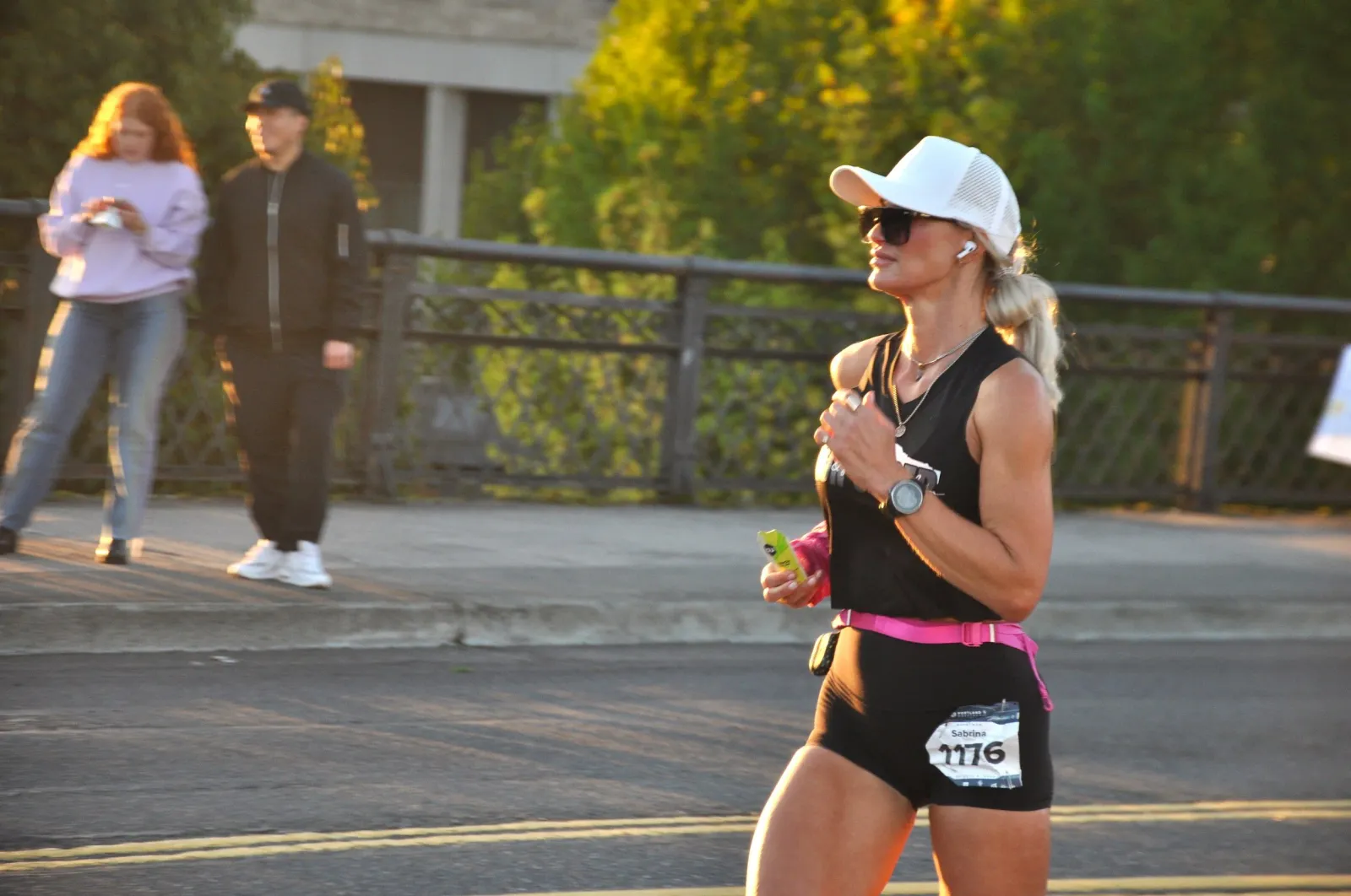 A female runner wearing a black tank top, black shorts with a pink belt, and a white cap runs on a road during a race. She is wearing sunglasses and earbuds, and has a race bib with the number 1176 on her shorts. Two other people are visible in the background.