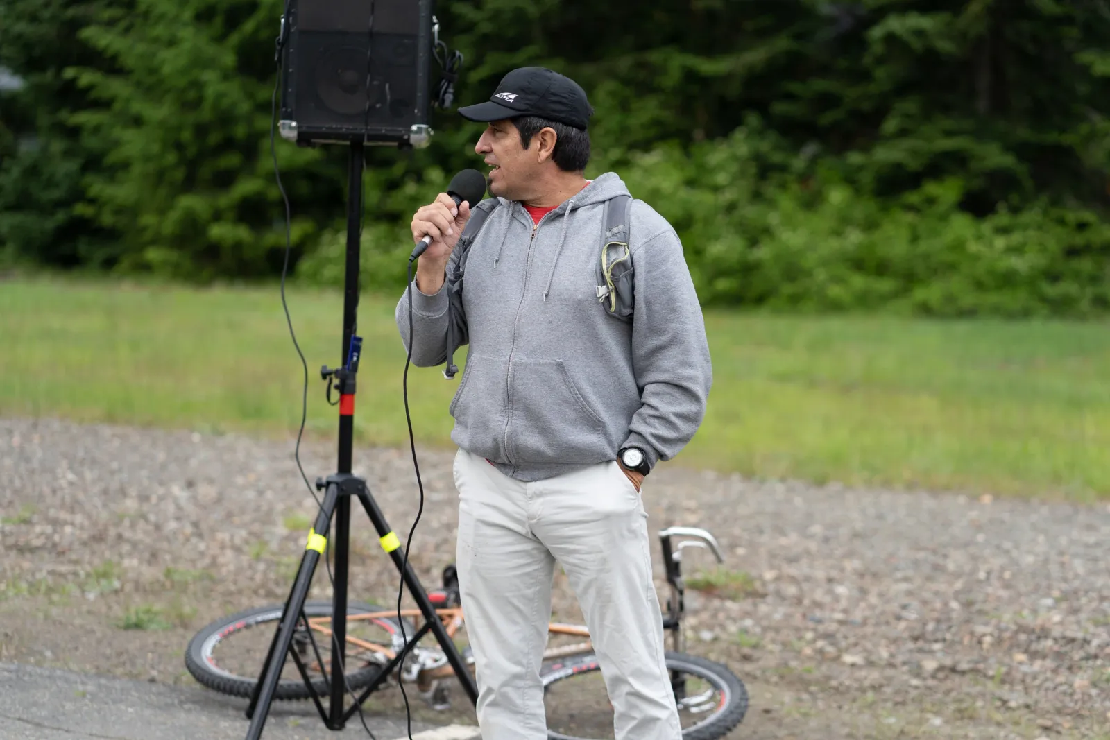 A man wearing a black baseball cap and a grey hoodie speaks into a microphone, standing next to a speaker on a stand with a bicycle lying on the ground behind him.