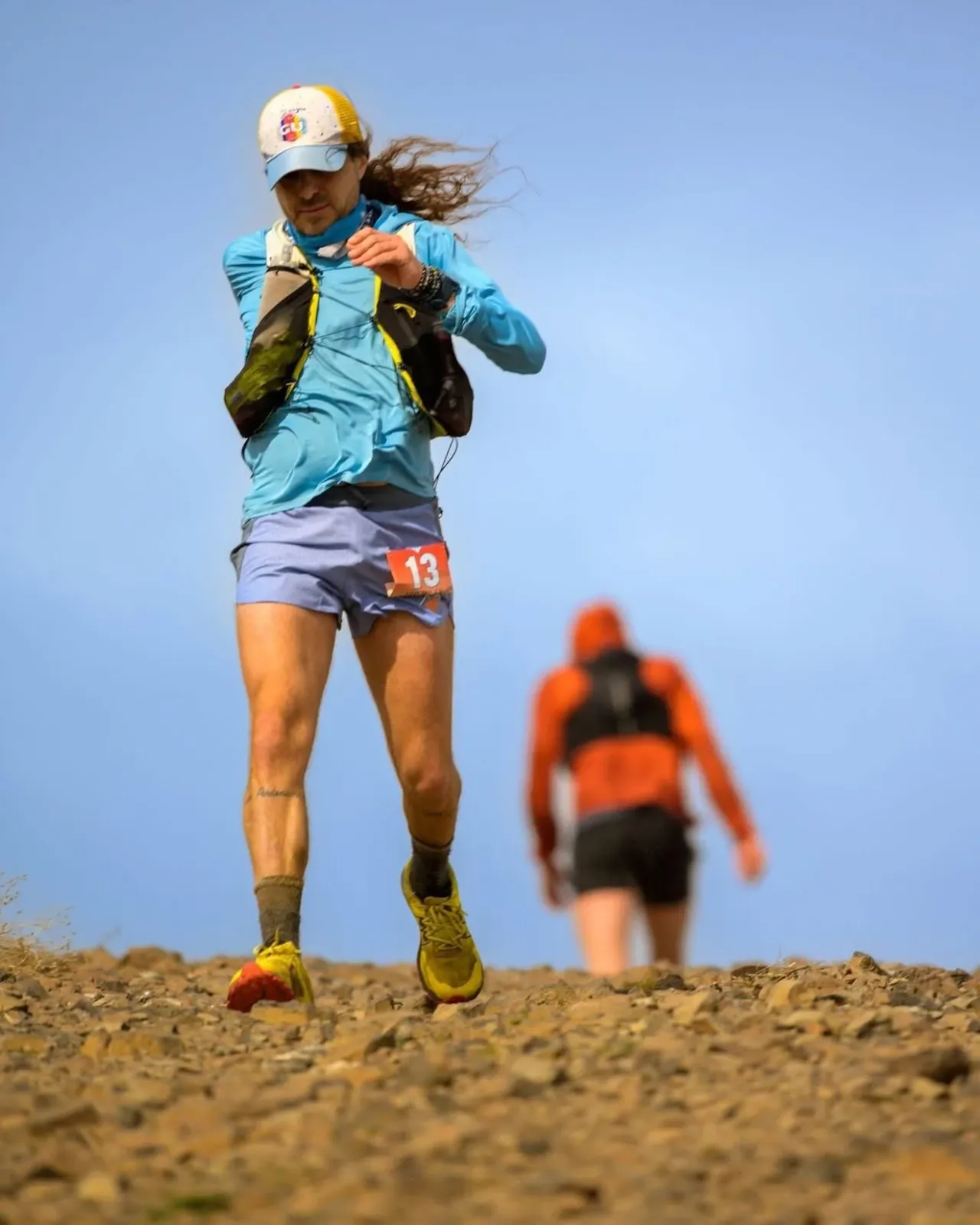 A runner with a bib number 13 on their shorts runs uphill on a rocky trail under a clear blue sky.