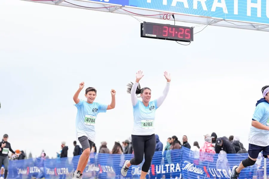 A young boy and a woman with their arms raised in celebration as they cross the finish line of a marathon, with a digital timer showing 34:25.