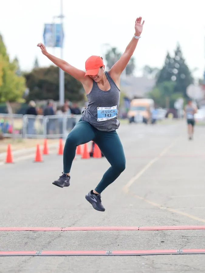 A female runner in mid-air, celebrating as she crosses the finish line, wearing an orange cap and grey tank top.