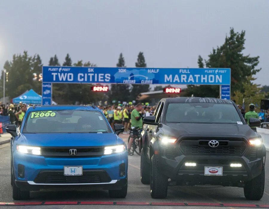 Start line of the Two Cities Marathon with runners gathered behind two vehicles and a large banner overhead.