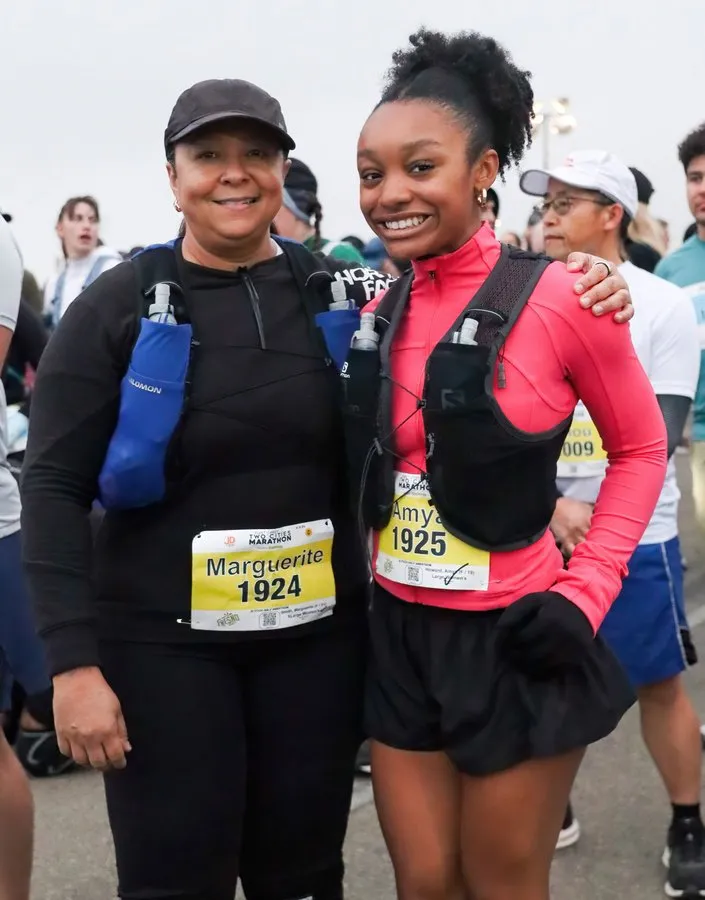 Two female runners, one older and one younger, smiling at the camera before a race. Both are wearing running gear and bibs.