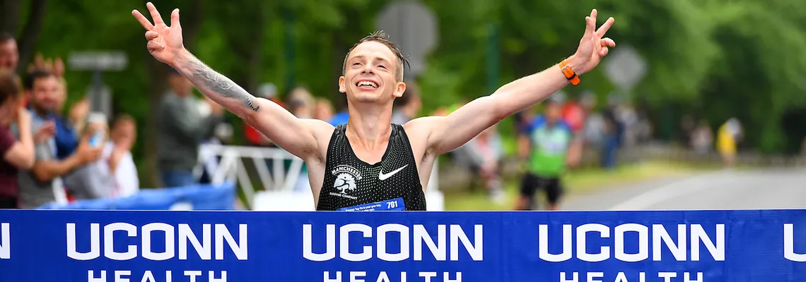 A male runner with arms raised in victory crosses the finish line, celebrating with a V-sign gesture, with the race banner visible.