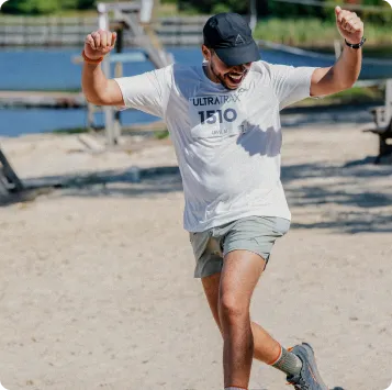 A male runner wearing a white Ultratrax t-shirt with the number 1510, grey shorts, and a black cap, pumps his fists in the air with a look of joy and excitement.