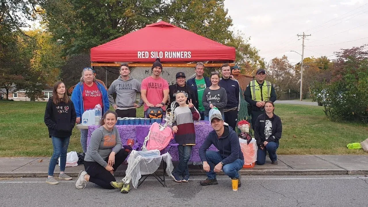 A group of people, some wearing 'Red Silo Runners' apparel, stand and pose around a red tent at an outdoor event, with Halloween decorations visible.