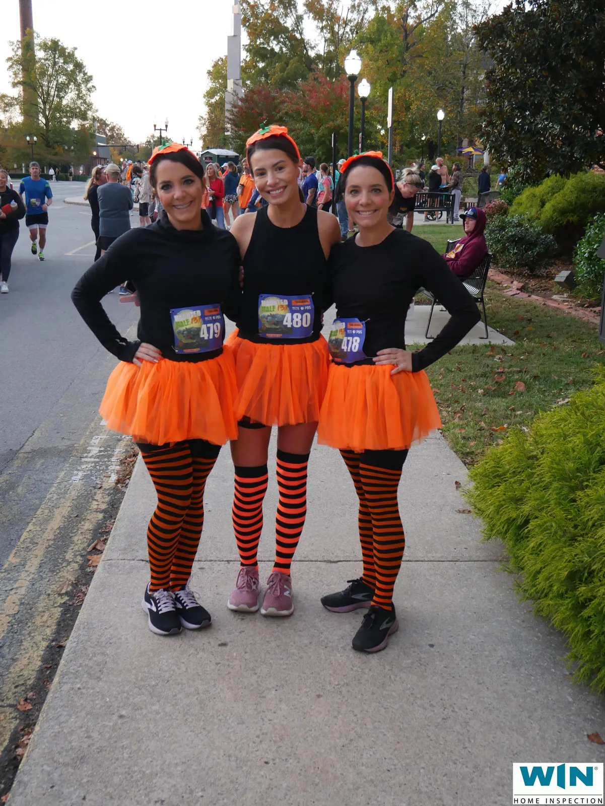 Three women in Halloween costumes, including orange tutus and striped socks, pose for a photo at a race.