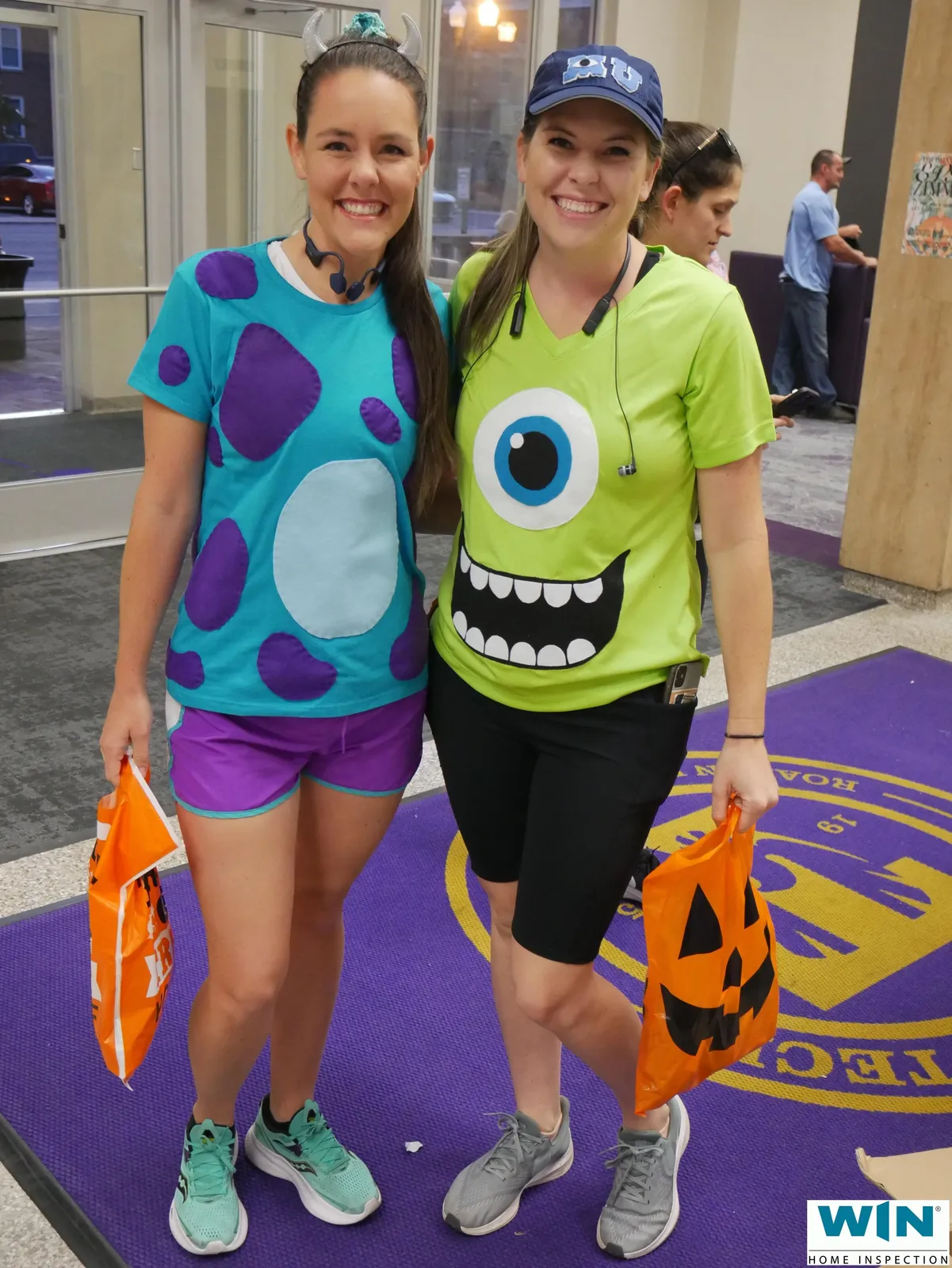 Two women in Monsters Inc. themed costumes smiling and holding Halloween bags at a race event.