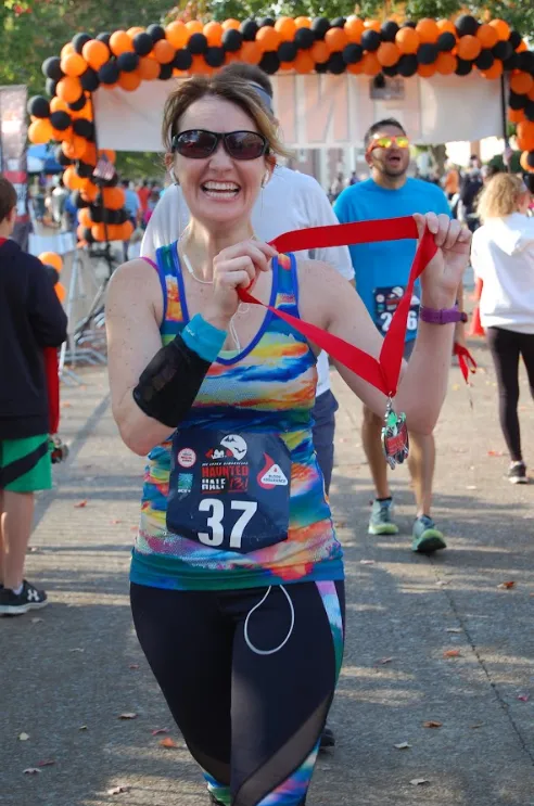 A smiling female runner holds up her medal after finishing the Upper Cumberland Haunted Half Marathon, with a festive balloon arch in the background.