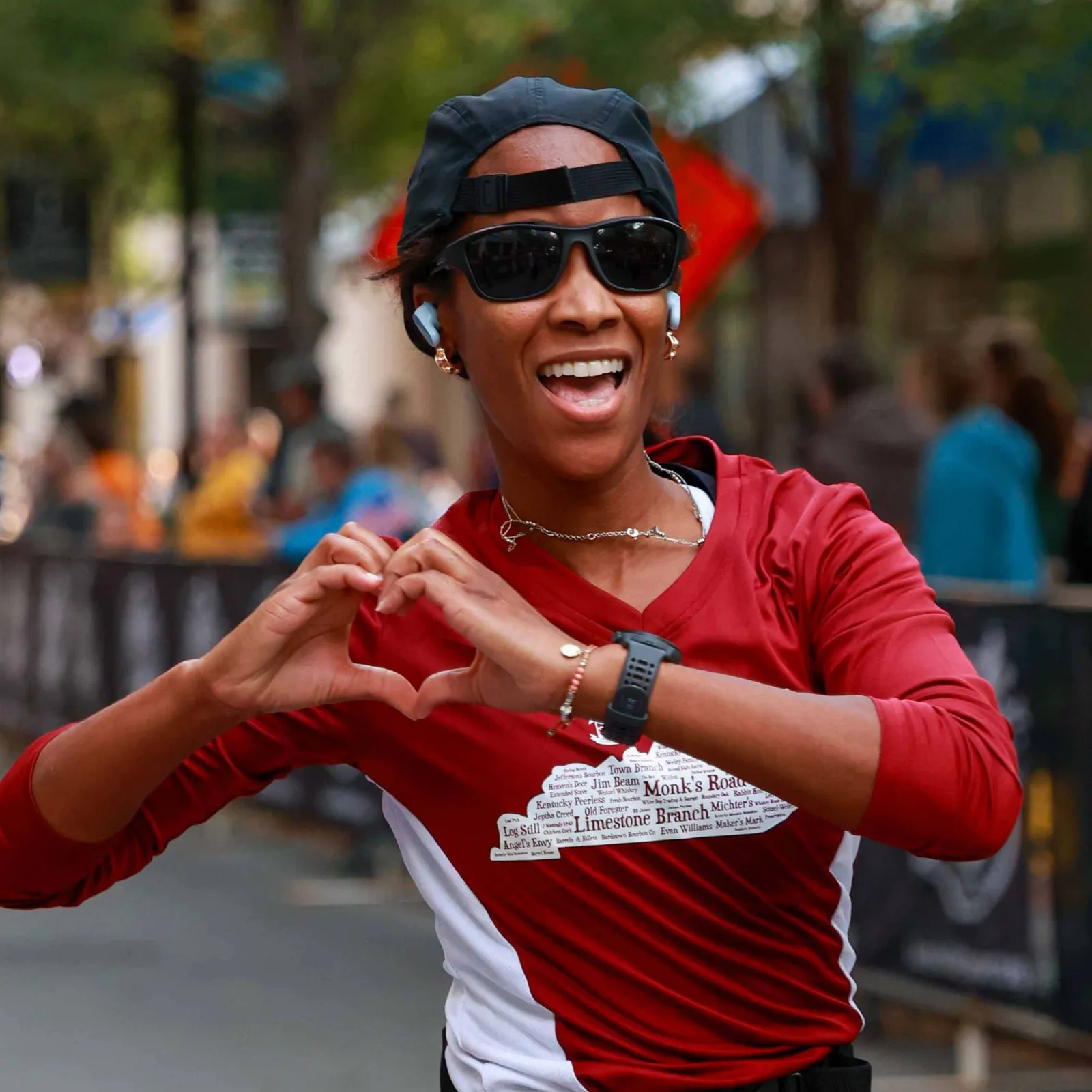 A female runner wearing a red shirt and a black baseball cap smiles and makes a heart shape with her hands during a race.