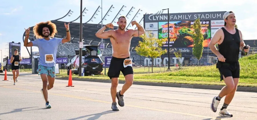 Three runners on a road with a stadium in the background. The runner in the middle is shirtless and flexing, while the runner on the left is smiling and making a rock-on gesture.