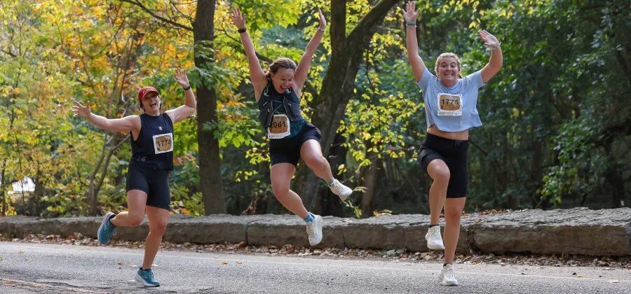 Three women runners joyfully jumping in the air during a race, with trees and fall foliage in the background.