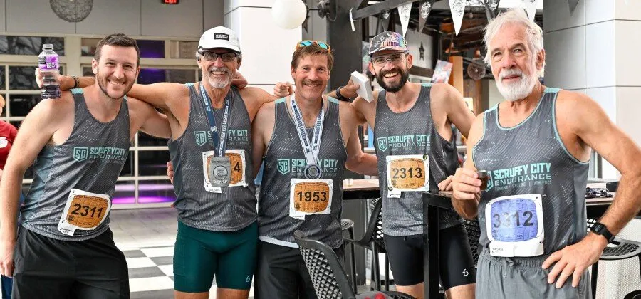 Five runners, all wearing similar grey tank tops with 'Scruffy City Endurance' branding and race bibs, pose together with medals around their necks, smiling and appearing to celebrate after a race.