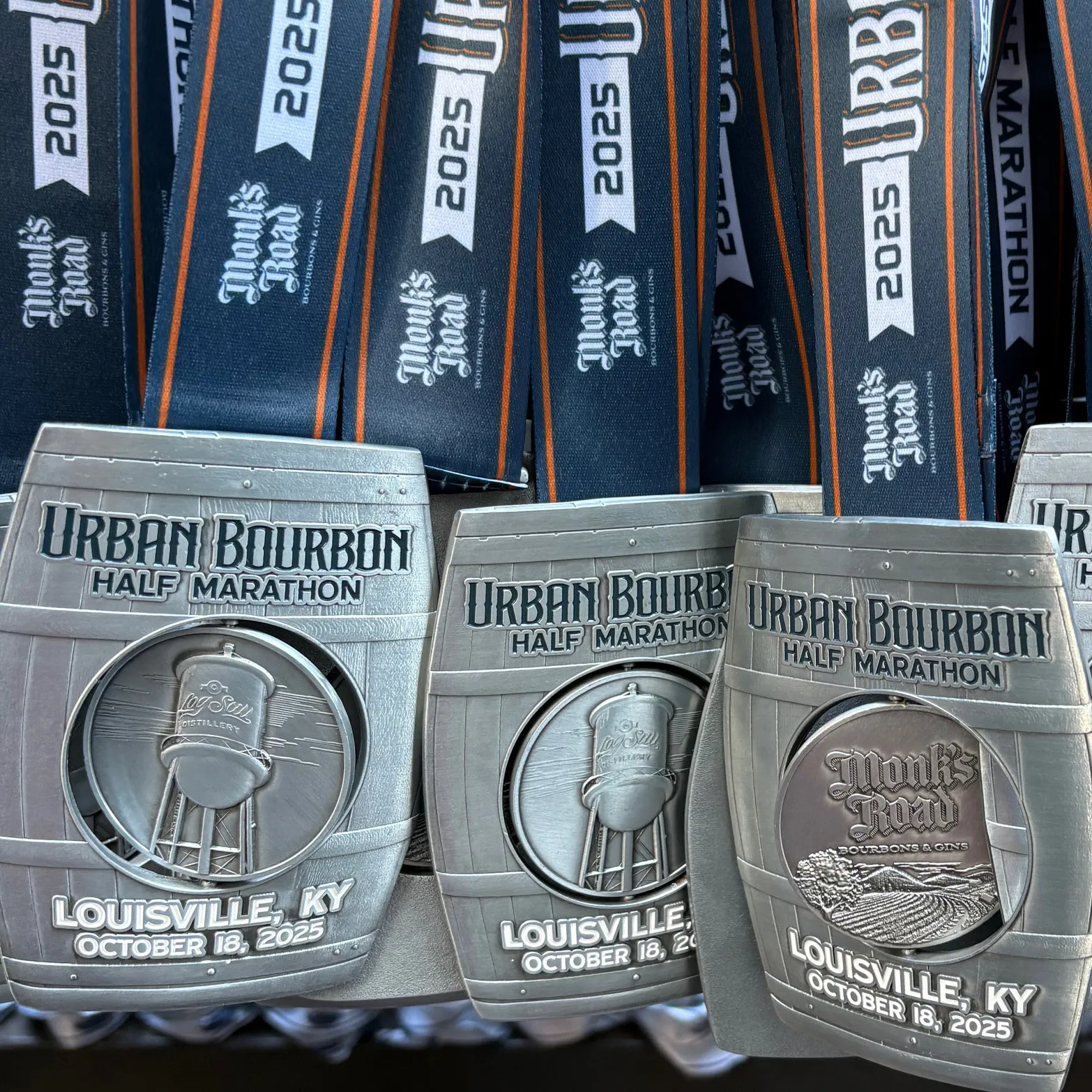 Close-up of three Urban Bourbon Half Marathon medals, shaped like barrels, with ribbons displaying 'Monk's Road' and '2025'.