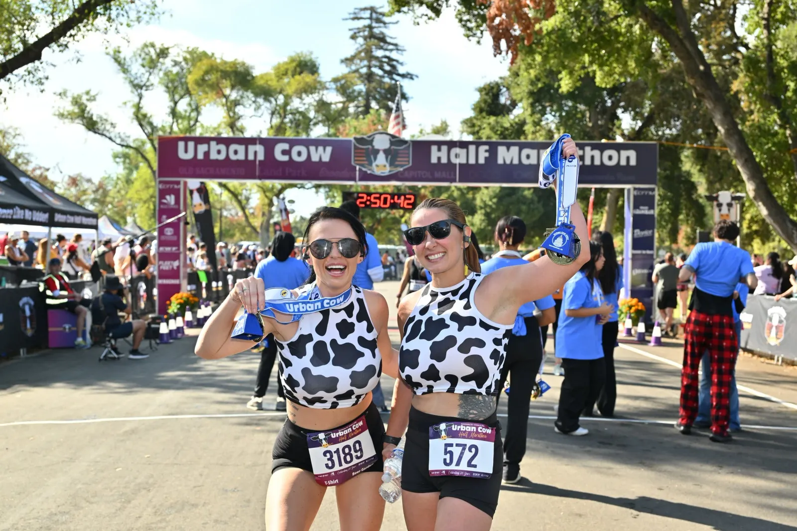 Two smiling women in cow-print tops celebrate at the finish line of the Urban Cow Half Marathon, holding up their medals.