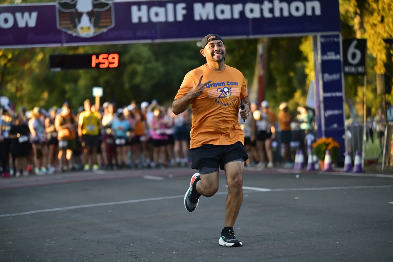 A male runner in an orange 'Urban Cow Half Marathon' shirt smiles and gives a peace sign as he crosses the finish line, with a crowd and race banner in the background.