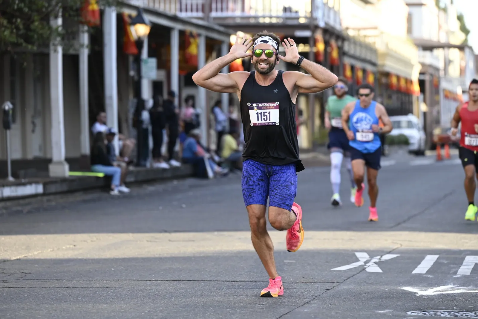 A male runner with his hands raised in excitement, wearing bib number 1151, runs down an urban street during the Urban Cow Half Marathon.