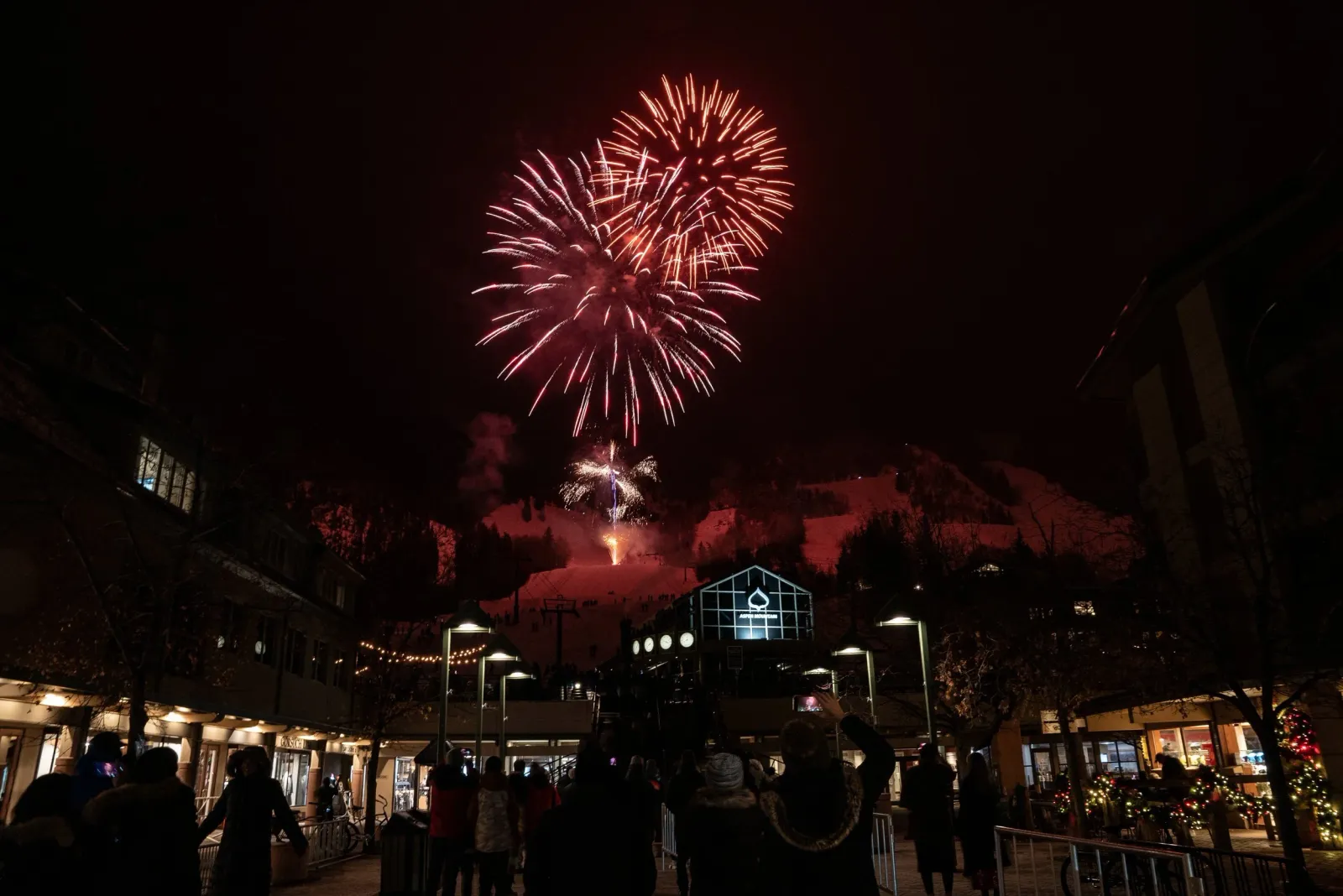 Fireworks explode in the night sky over a snowy mountain resort town, with a crowd of people watching below.