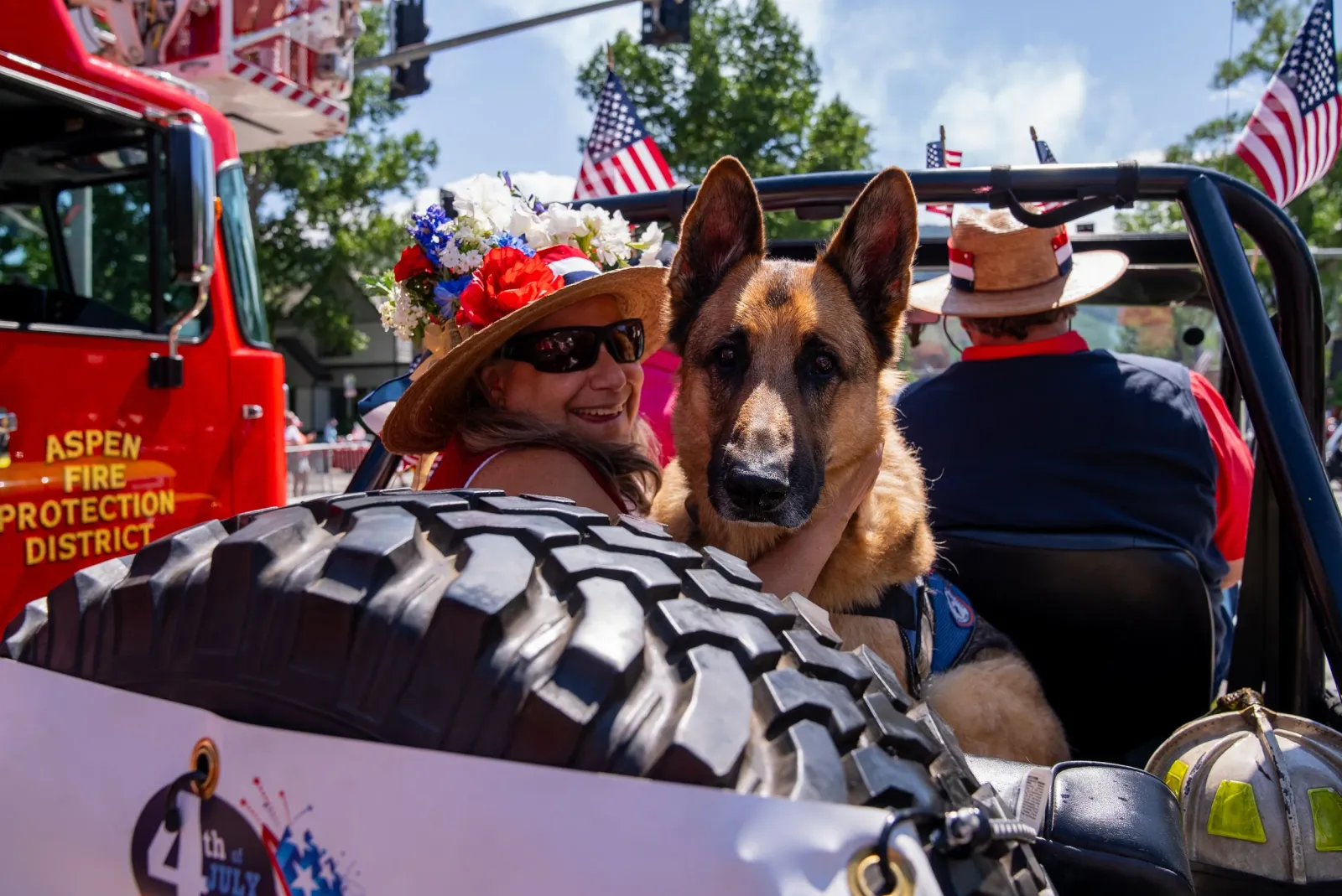 A woman in a straw hat decorated with flowers smiles next to a German Shepherd in a parade float, with a fire truck in the background.
