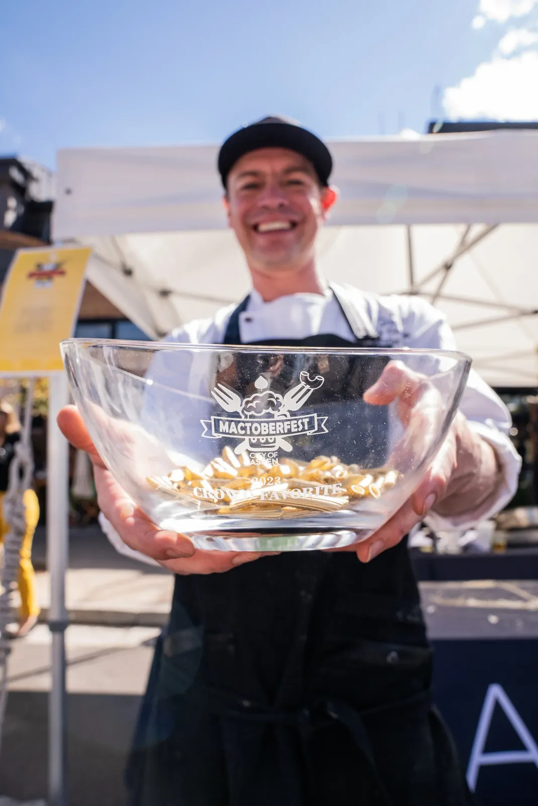 A smiling man in a chef's coat and apron holds a glass bowl filled with small golden objects, with the text 'Mactoberfest City of Aspen 2023 Crowd Favorite' visible on the bowl.