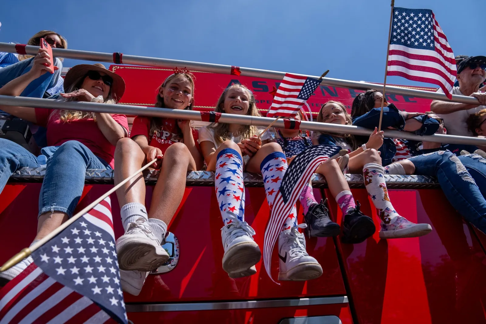 A group of people, including children, sitting on the side of a red vehicle, waving American flags and smiling.