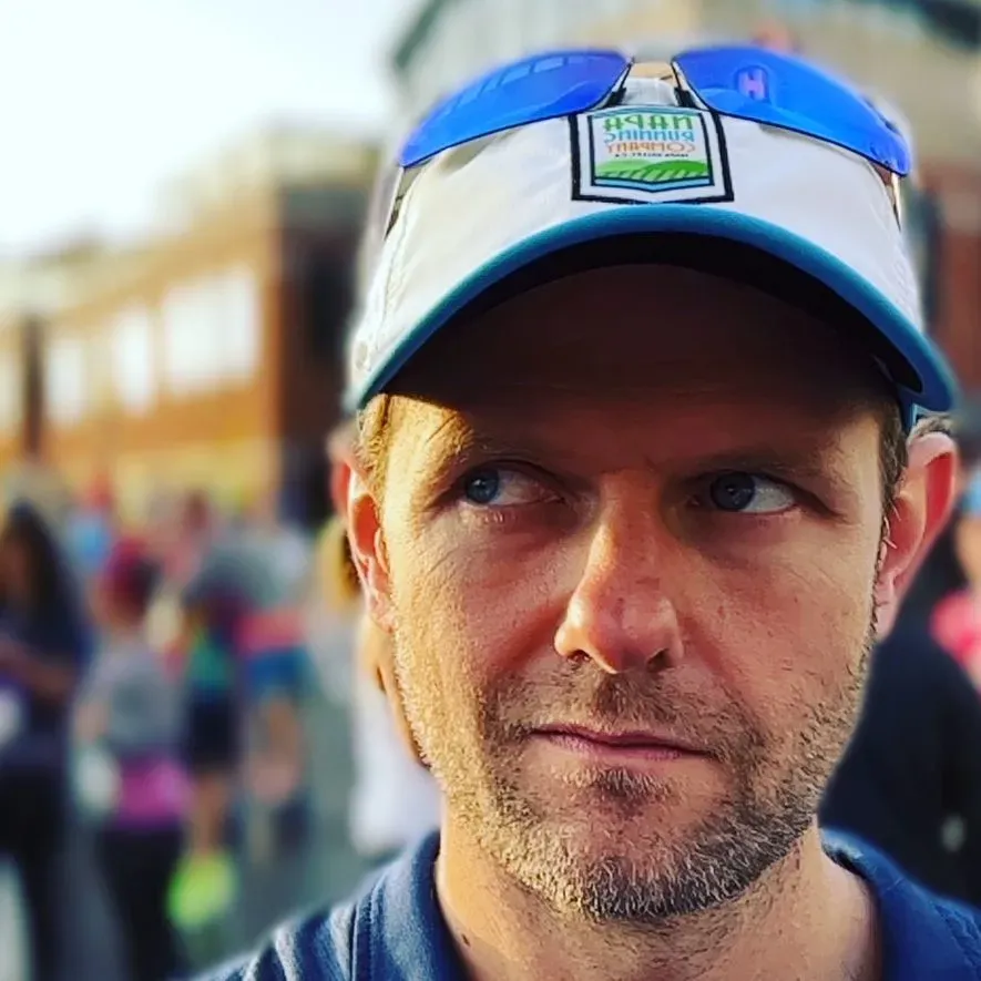 Close-up of a male runner wearing a blue polo shirt and a white running cap with sunglasses on top, looking to the side with a crowd blurred in the background.