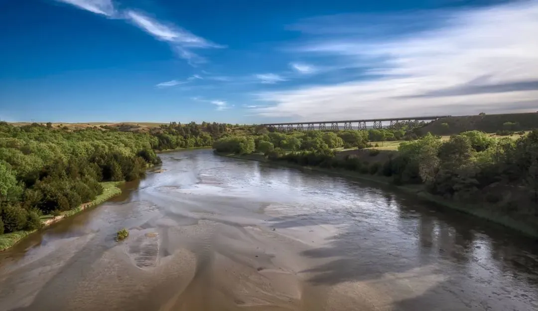 A wide river flows through a green landscape with a large bridge in the distance under a blue sky with wispy clouds.