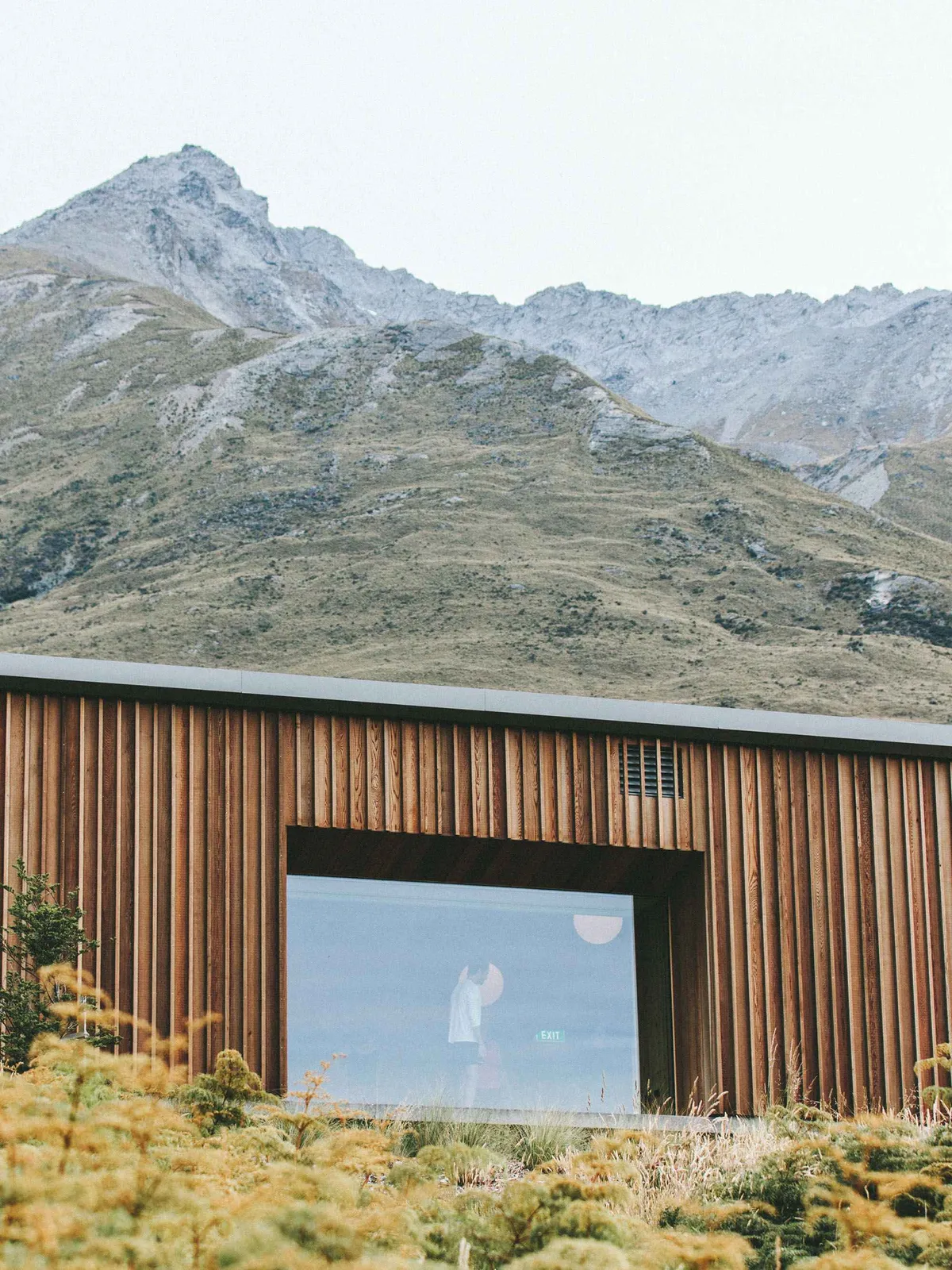 Scenic view of a mountain range behind a modern wooden building with a large window reflecting a person.