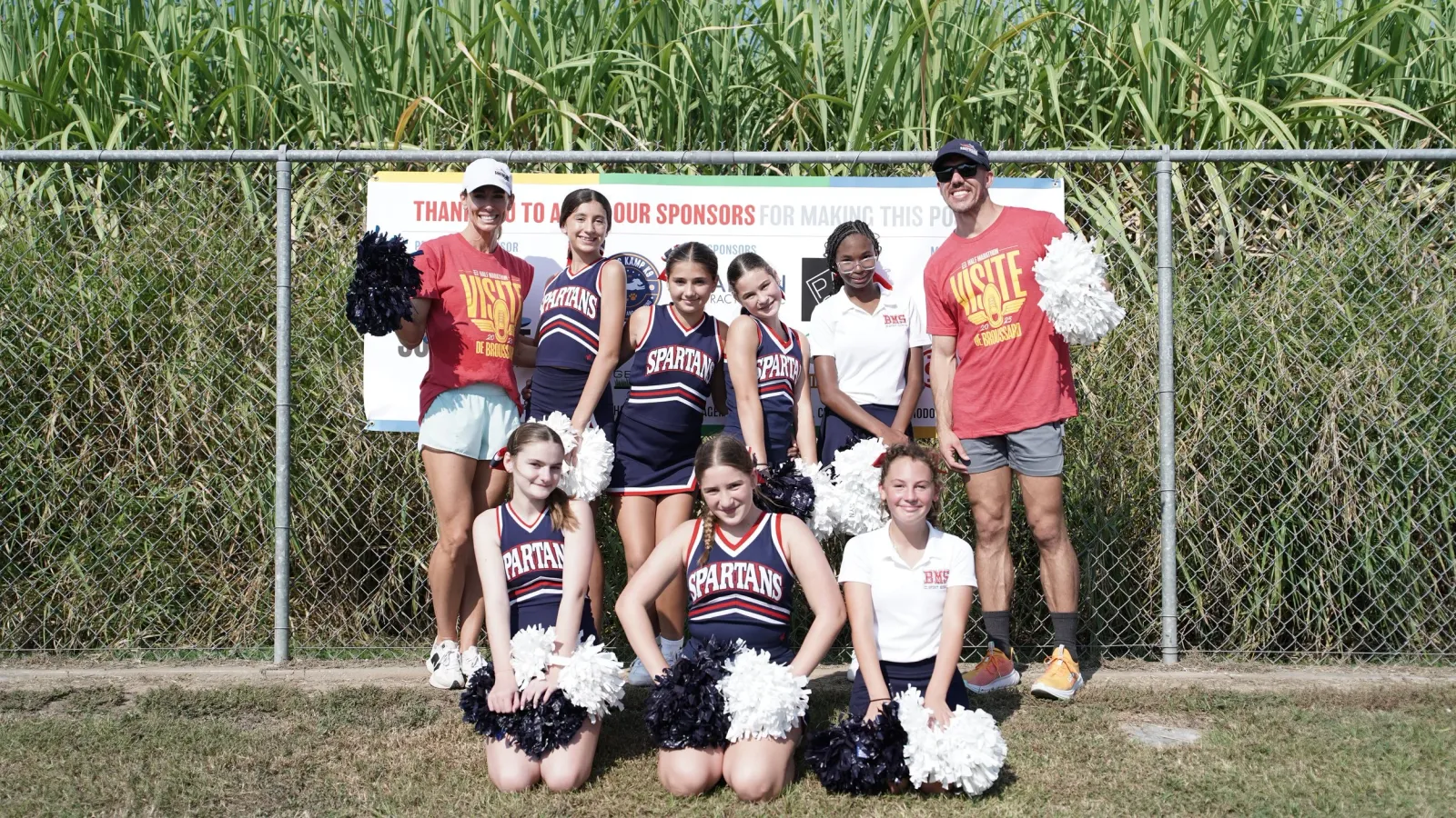 A group of cheerleaders and two adults pose for a photo in front of a fence and tall grass, with a sponsor banner in the background.