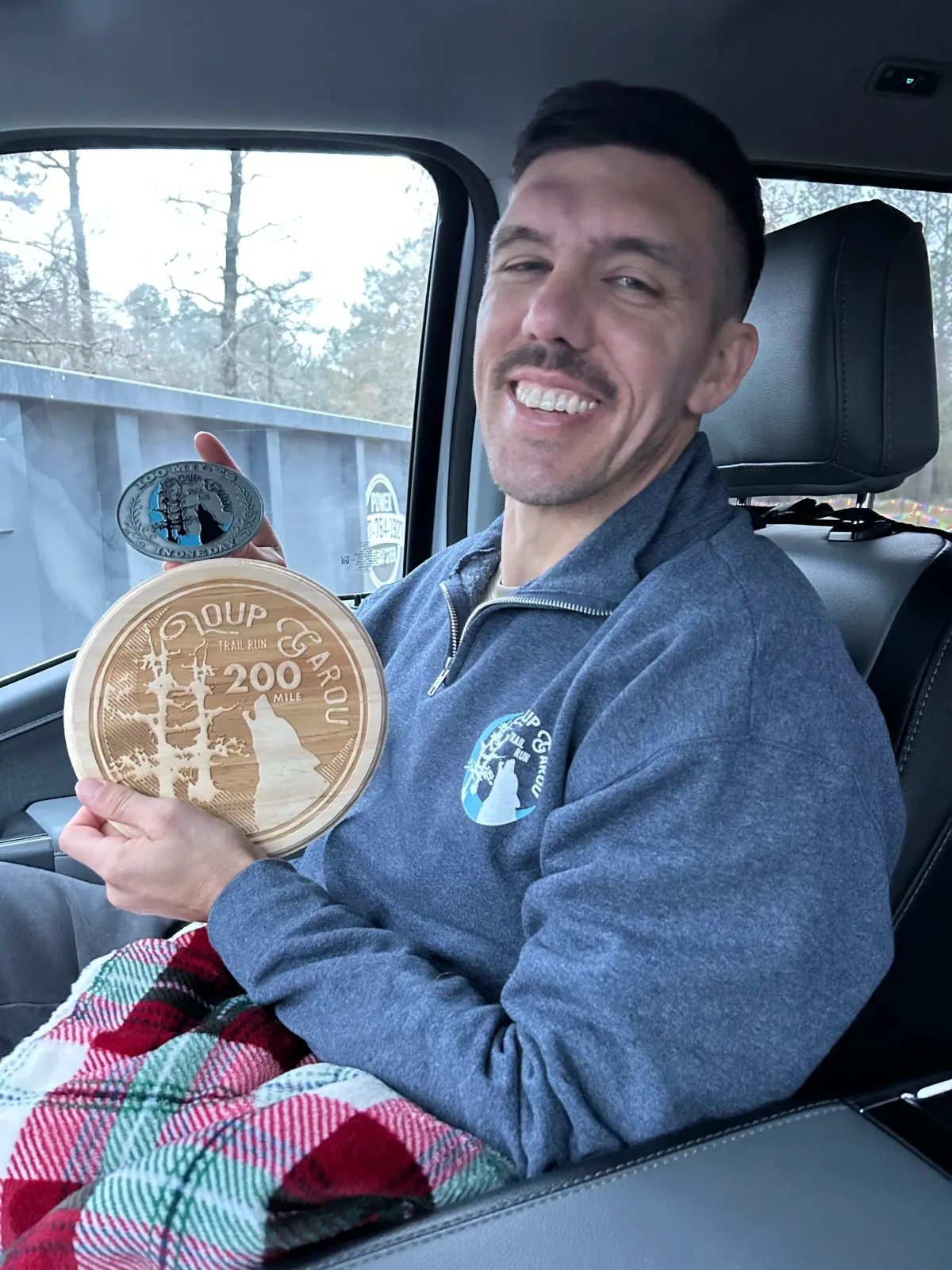 A smiling man sits in a car, holding a race medal and a wooden plaque that says "200 MILE TRAIL RUN".