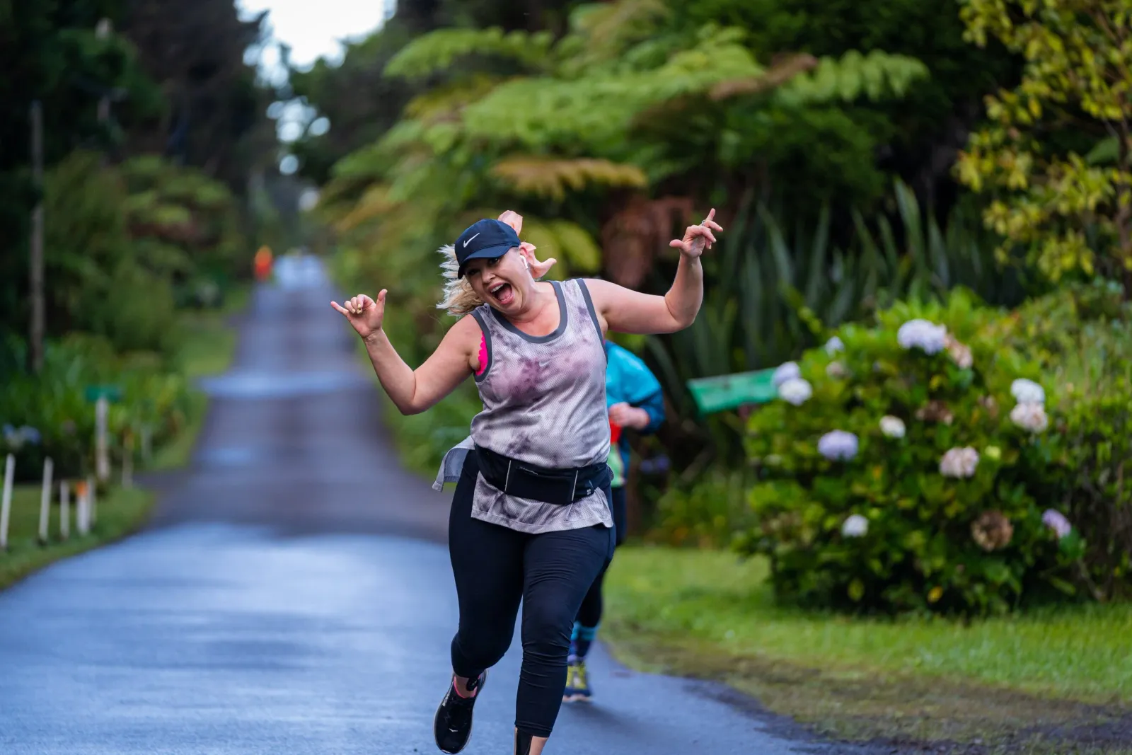 A female runner with a big smile and her hands in a 'shaka' gesture runs on a road lined with lush green foliage.