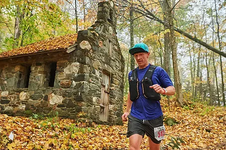 A male runner with a blue shirt and black vest runs on a trail past a stone building, with autumn leaves on the ground and trees in the background.