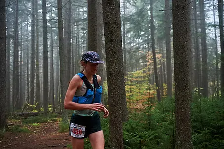 A female runner with a blue hydration vest runs on a forest trail, wearing a cap and shorts. A race bib is visible on her shorts.