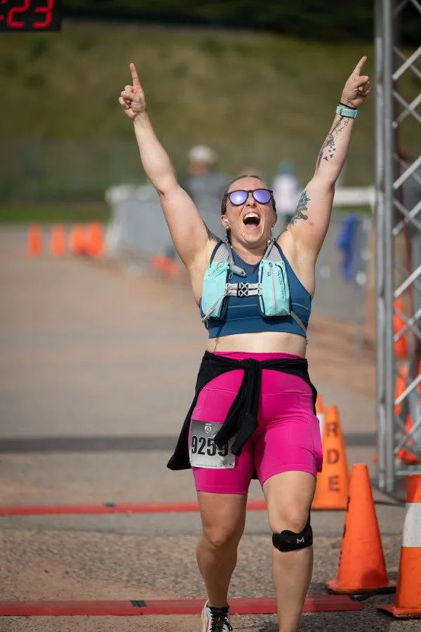 A female runner with arms raised in celebration crosses the finish line, wearing pink shorts and a blue sports bra.