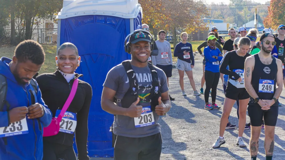 A group of runners in the foreground and background, some smiling and giving thumbs up, at the start of a race.