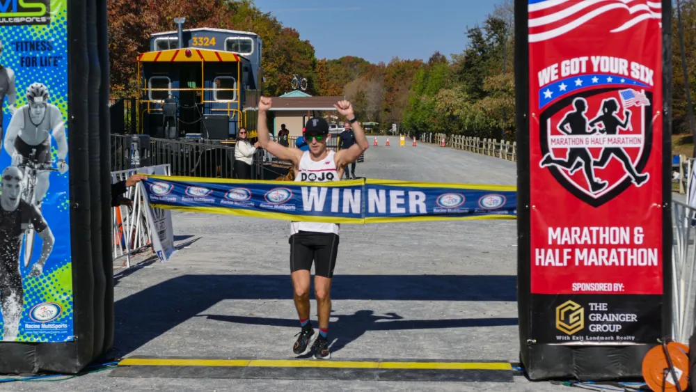 A male runner crosses the finish line with his arms raised in celebration, breaking a 'WINNER' banner.