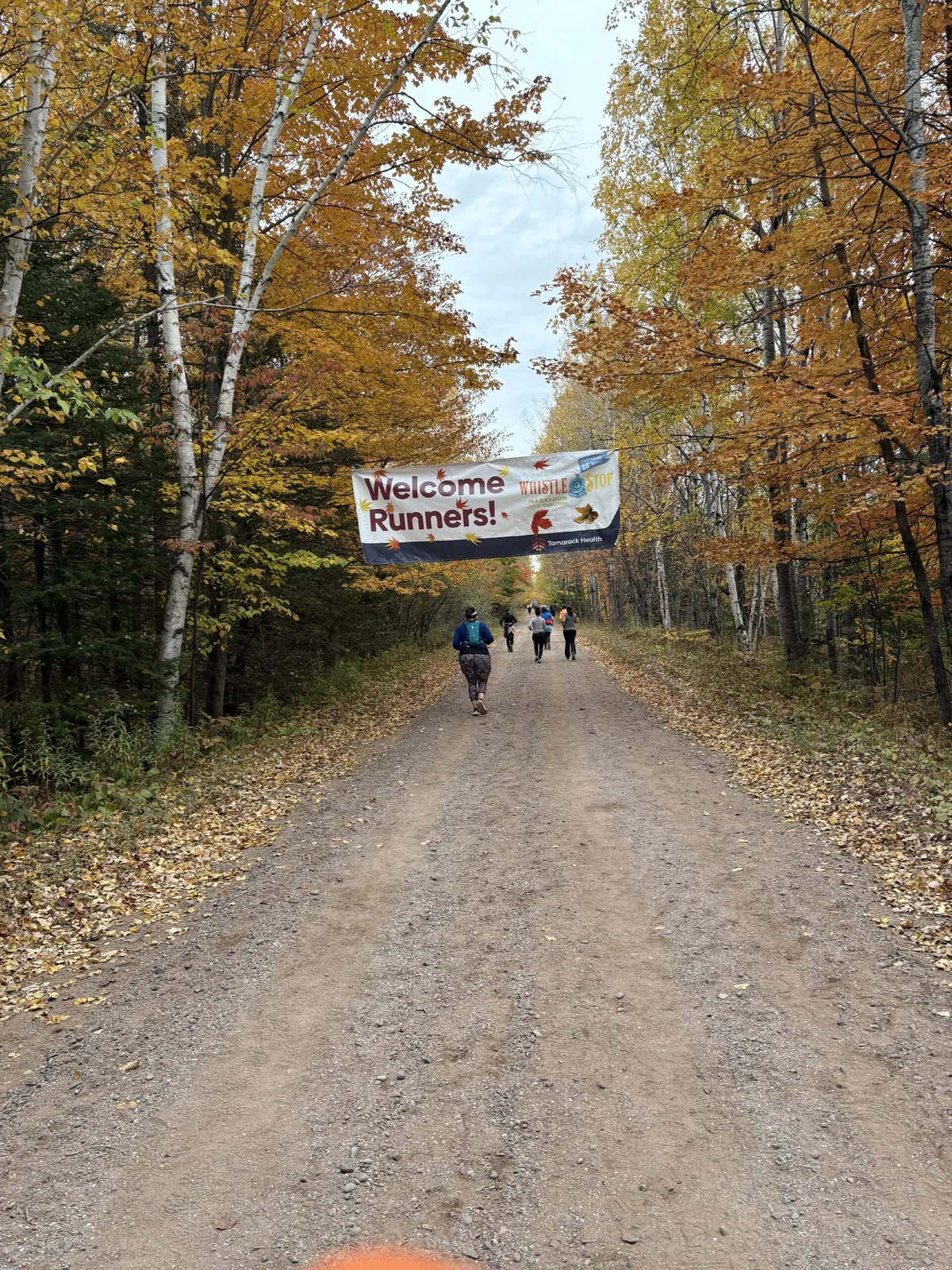 Runners on a gravel road through a forest with autumn foliage, passing under a banner that reads 'Welcome Runners!'