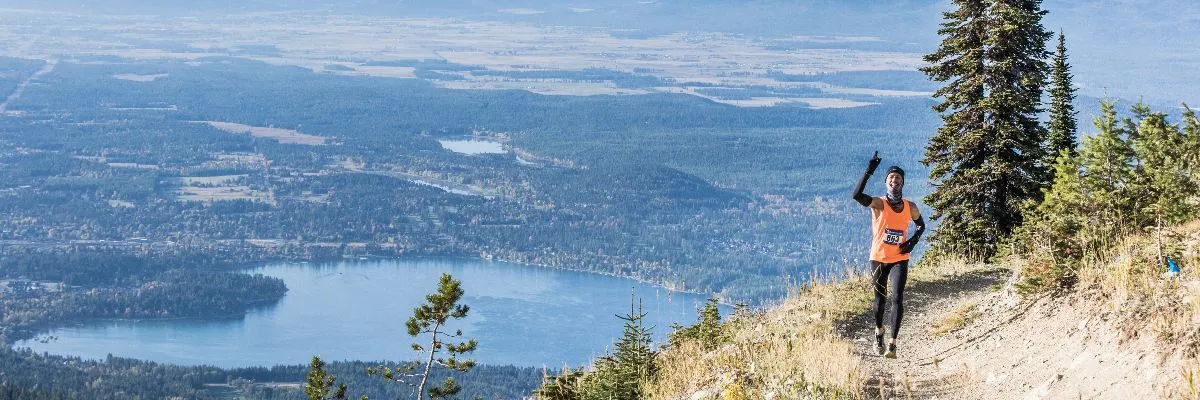 A runner in an orange tank top and black leggings smiles and points upwards while running on a mountain trail with a scenic lake and valley in the background.